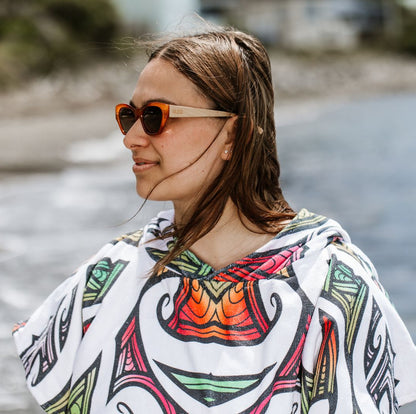 Woman at the beach wearing burnt orange coloured plastic frame sunglasses with brown polarized lenses and wooden arms. She also has a white towel hoodie on with multi coloured kory pattern. 