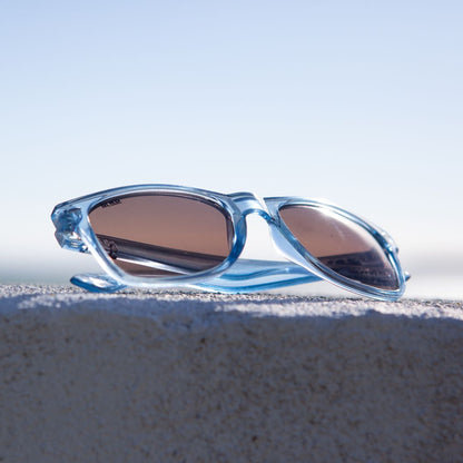 Denim blue coloured plastic framed sunglasses with brown polarized lenses placed on a stone wall with blurred sky in the background.