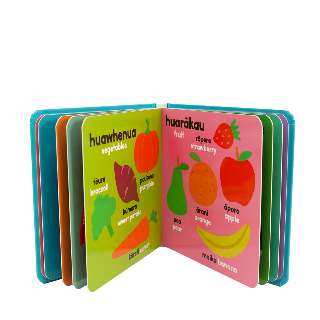 A children's learning book shown open on a white background, with colorful illustrated pages featuring labels such as huawhenua vegetables, huarākau fruit, strawberry, broccoli, pumpkin, sweet potato, pea, orange, apple, and banana and the Te Reo Maori words for each item.