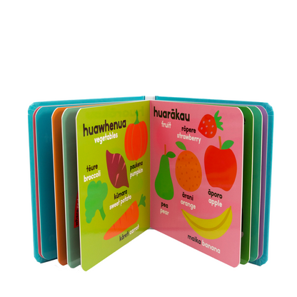 A children's learning book shown open on a white background, with colorful illustrated pages featuring labels such as huawhenua vegetables, huarākau fruit, strawberry, broccoli, pumpkin, sweet potato, pea, orange, apple, and banana and the Te Reo Maori words for each item.