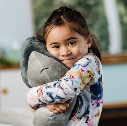 A young girl with dark hair is hugging a large gray plush Hectors Dolphin toy in a brightly lit room. She is wearing a colorful long-sleeve top with a blue, pink, and white floral bunny pattern.