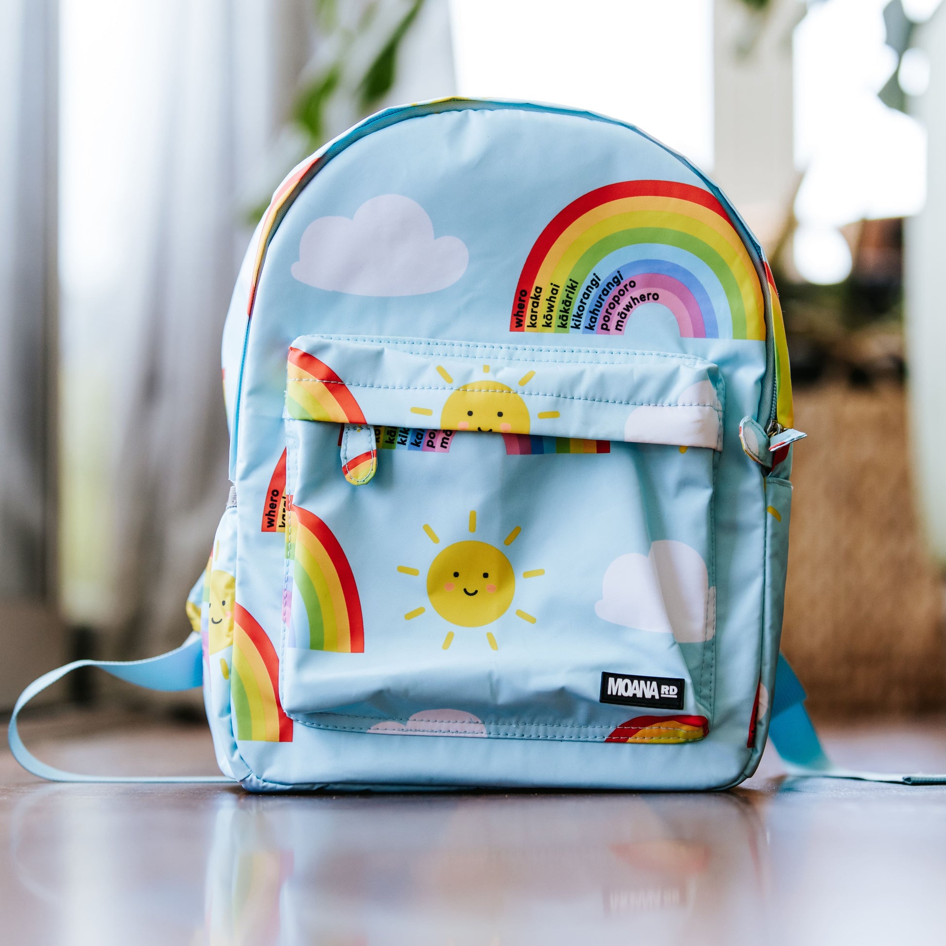 Light blue kids backpack with yellow smily face sun pictures and rainbows with te reo colours.