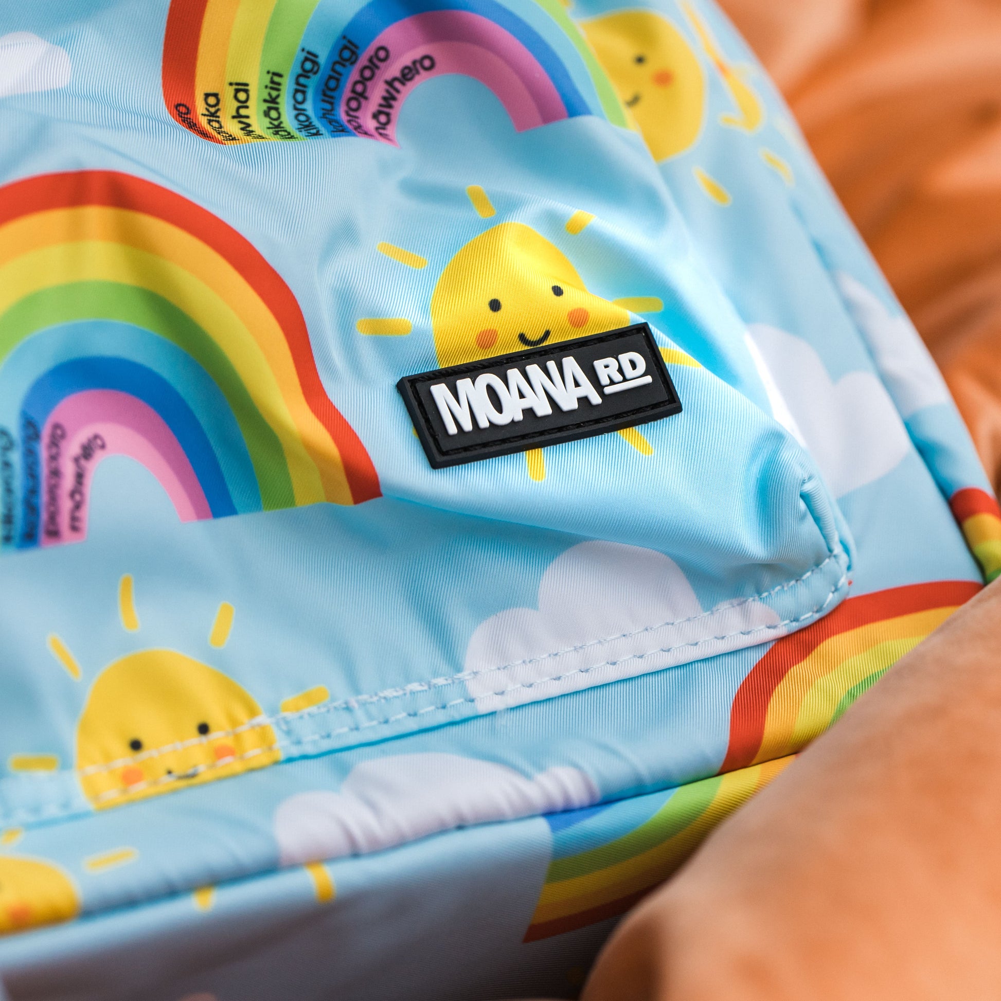 Corner of a Light blue kids backpack with yellow smily face sun pictures and rainbows with te reo colours. Showing the Moana RD label on the pocket.