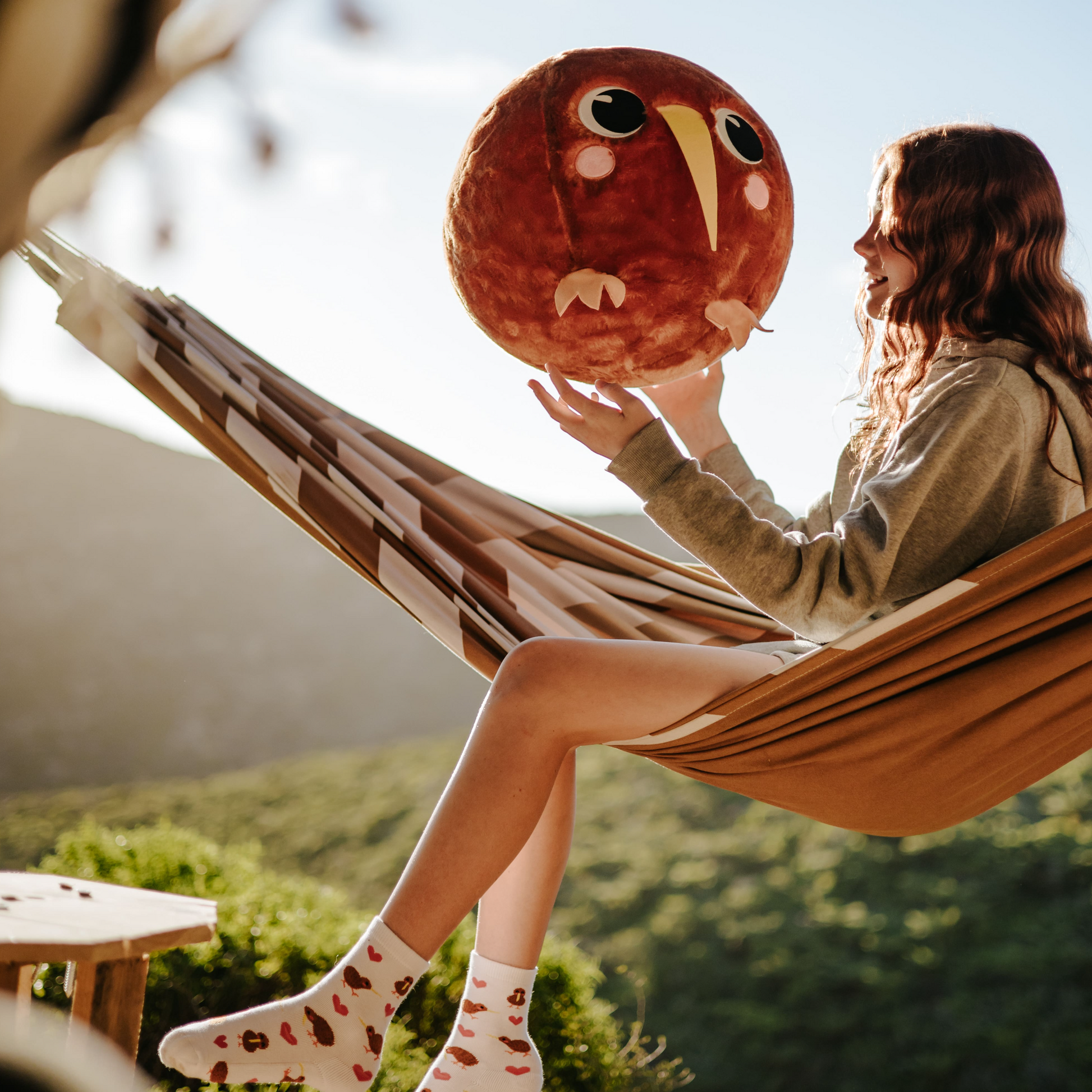Young girl sitting in a brown and white hammock holding onto an inflatable brown plush kiwi bird ball.