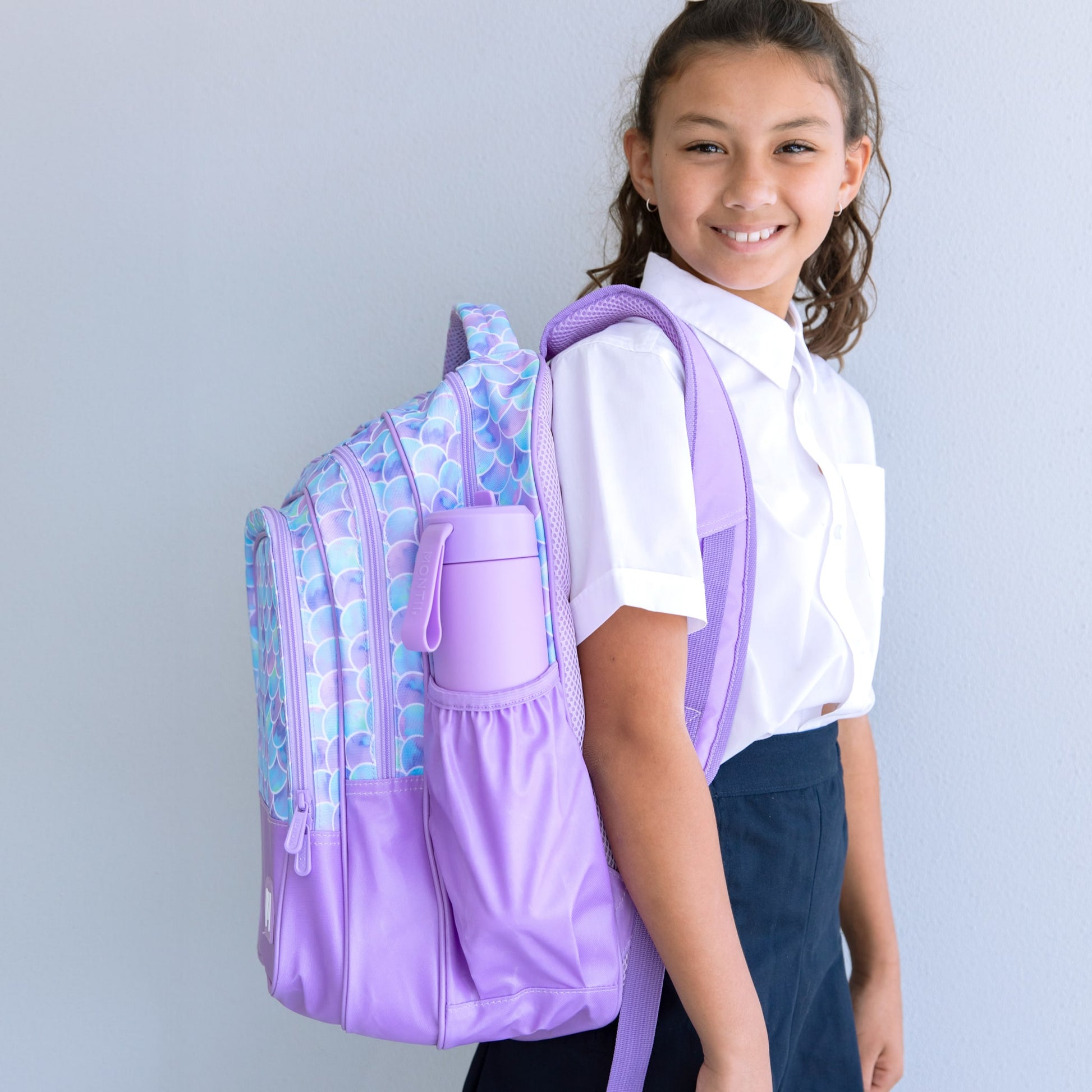 Young girl standing side on with a backpack over her right shoulder. The backpack is purple with a blue and purple mermaid scale print and has a purple drink bottle in the side pocket.