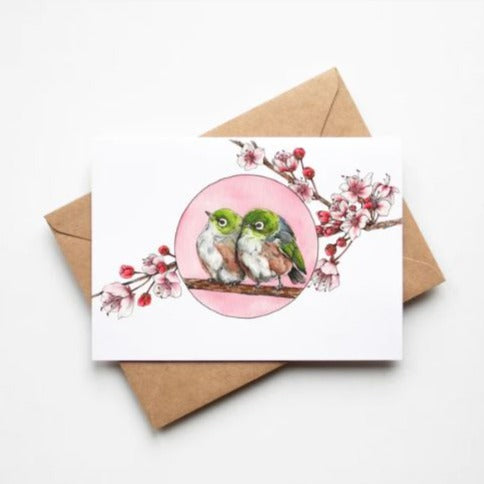 Greeting card by artist Leah Ingram featuring 2 Silvereye birds nestled together on a branch surrounded by pink blossoms.