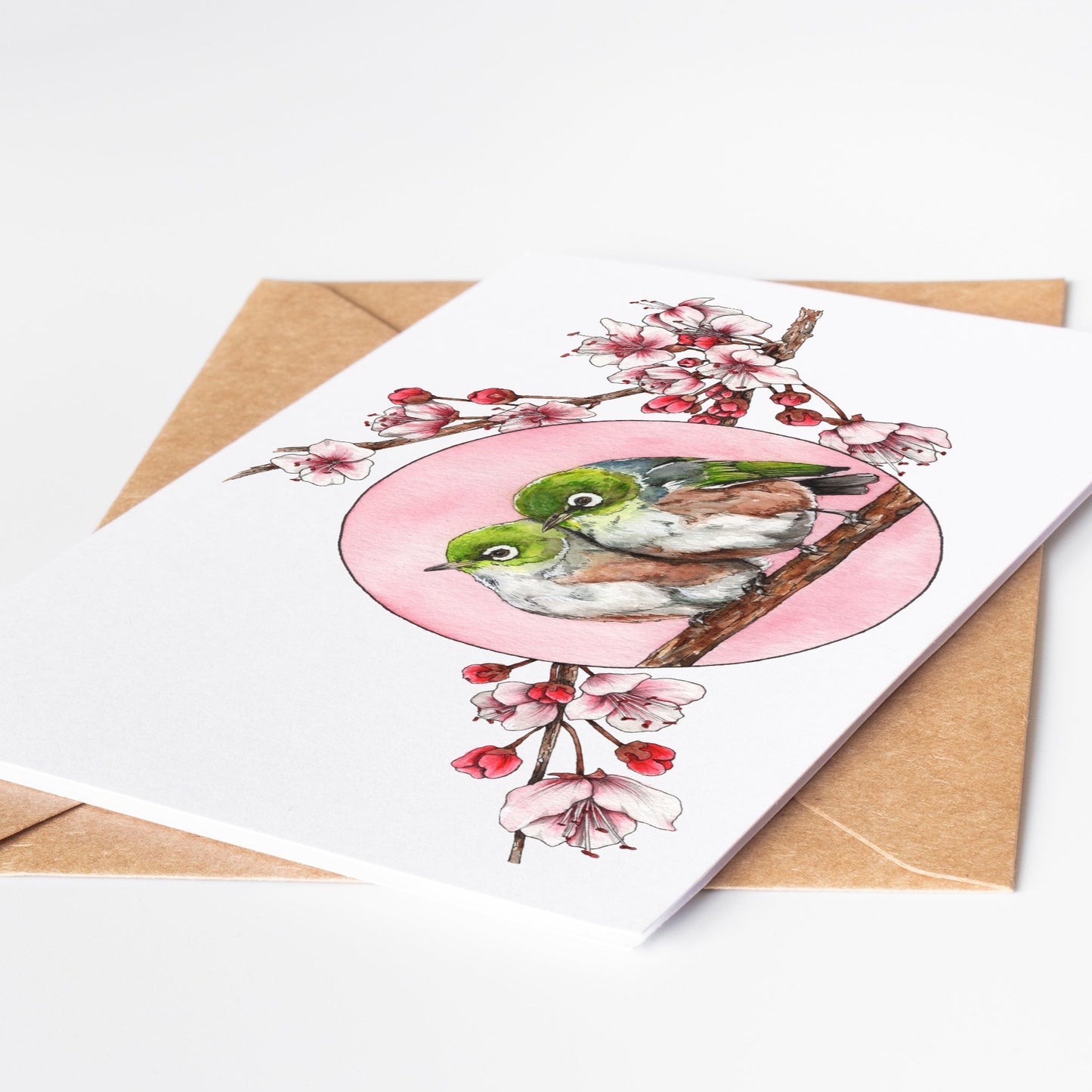 Greeting card by artist Leah Ingram featuring 2 Silvereye birds nestled together on a branch surrounded by pink blossoms.