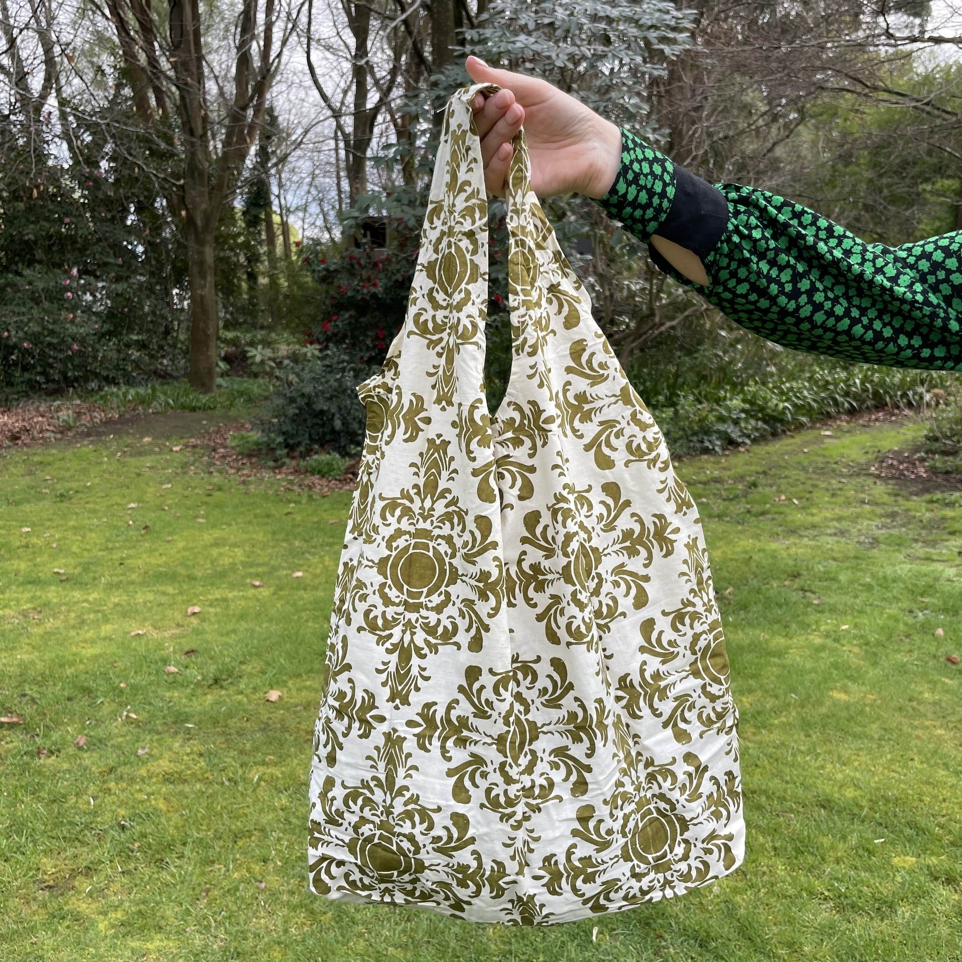 Womans hand holding a damask patterned olive green and white cotton tote bag with a lush green garden in the background.