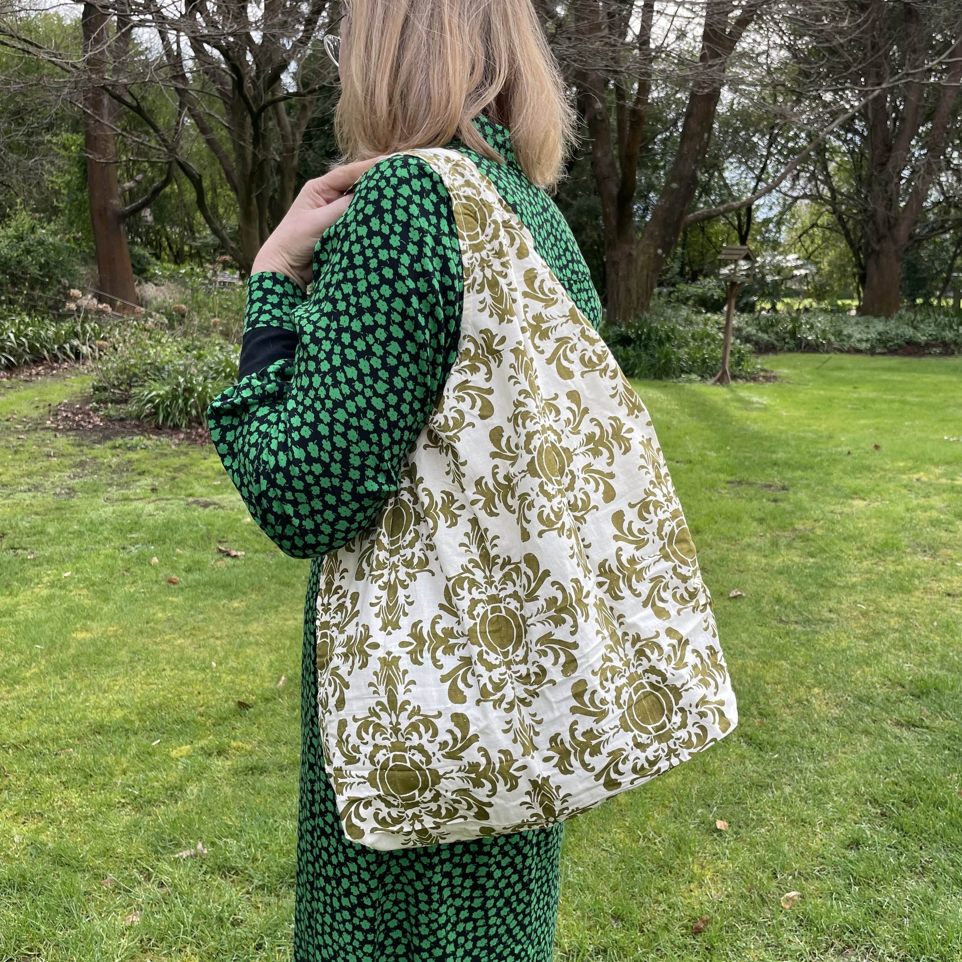Woman standing in a garden in a black and green dress with her back to the camera. She has a olive green and white damask patterned reusable cotton tote bag over her shoulder.
