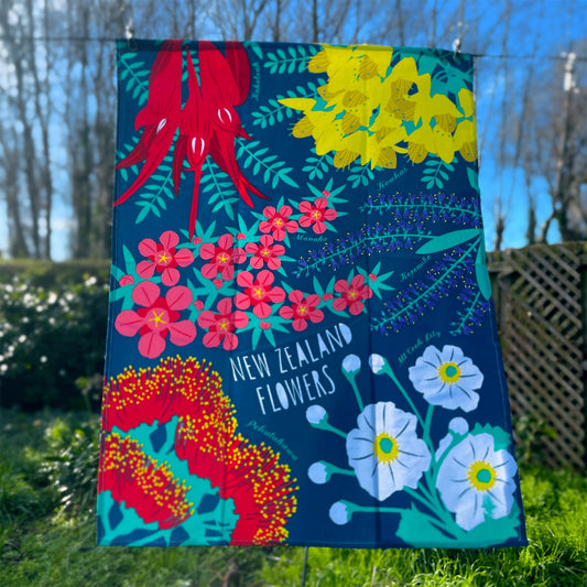 Dark teal blue tea towel with bright NZ flowers print hanging on a clothes line.