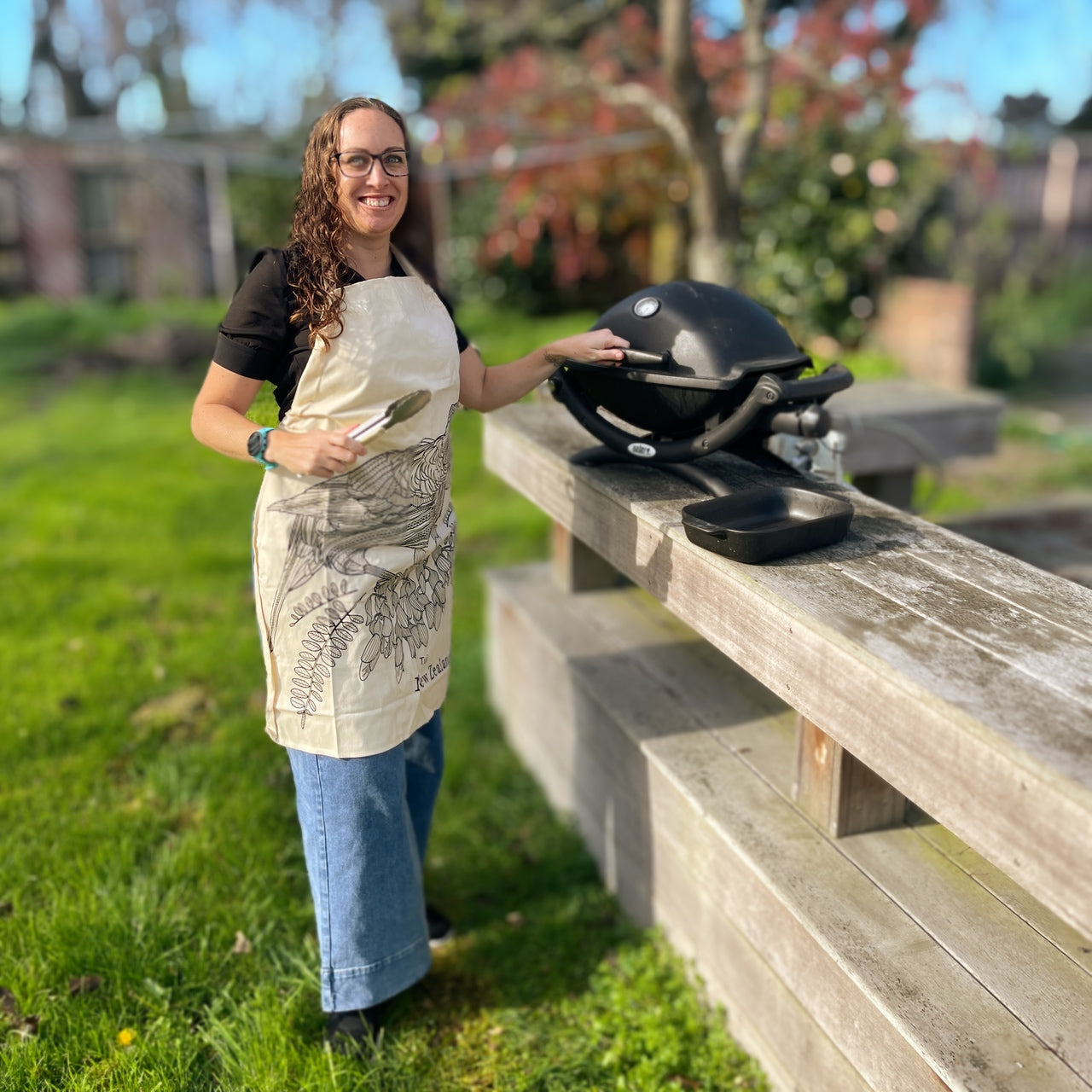 Woman wearing calico apron with black sketch of a Tui bird and kowhai flowers.