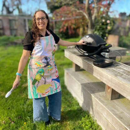 Woman wearing a peach coloured apron with bold and colourful NZ birds printed on it.