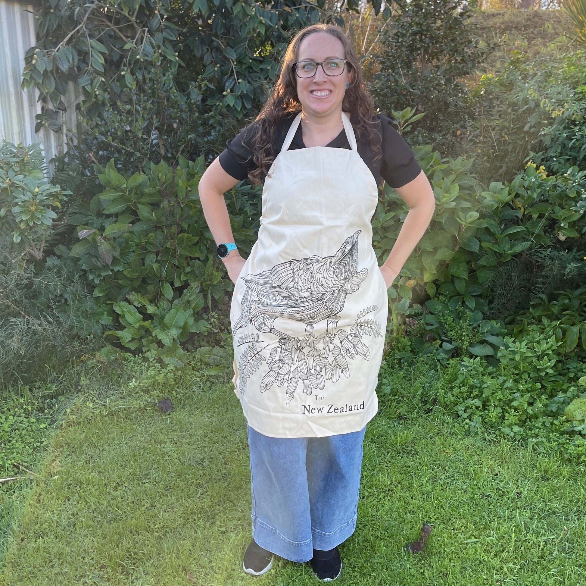 Woman wearing calico apron with black sketch of a Tui bird and kowhai flowers.