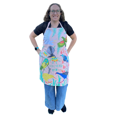 Woman wearing a peach coloured apron with bold and colourful NZ birds printed on it.