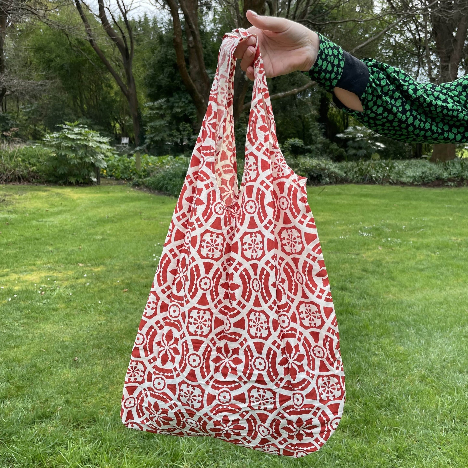 Womans hand holding a patterned red and white cotton tote bag with a lush green garden in the background.