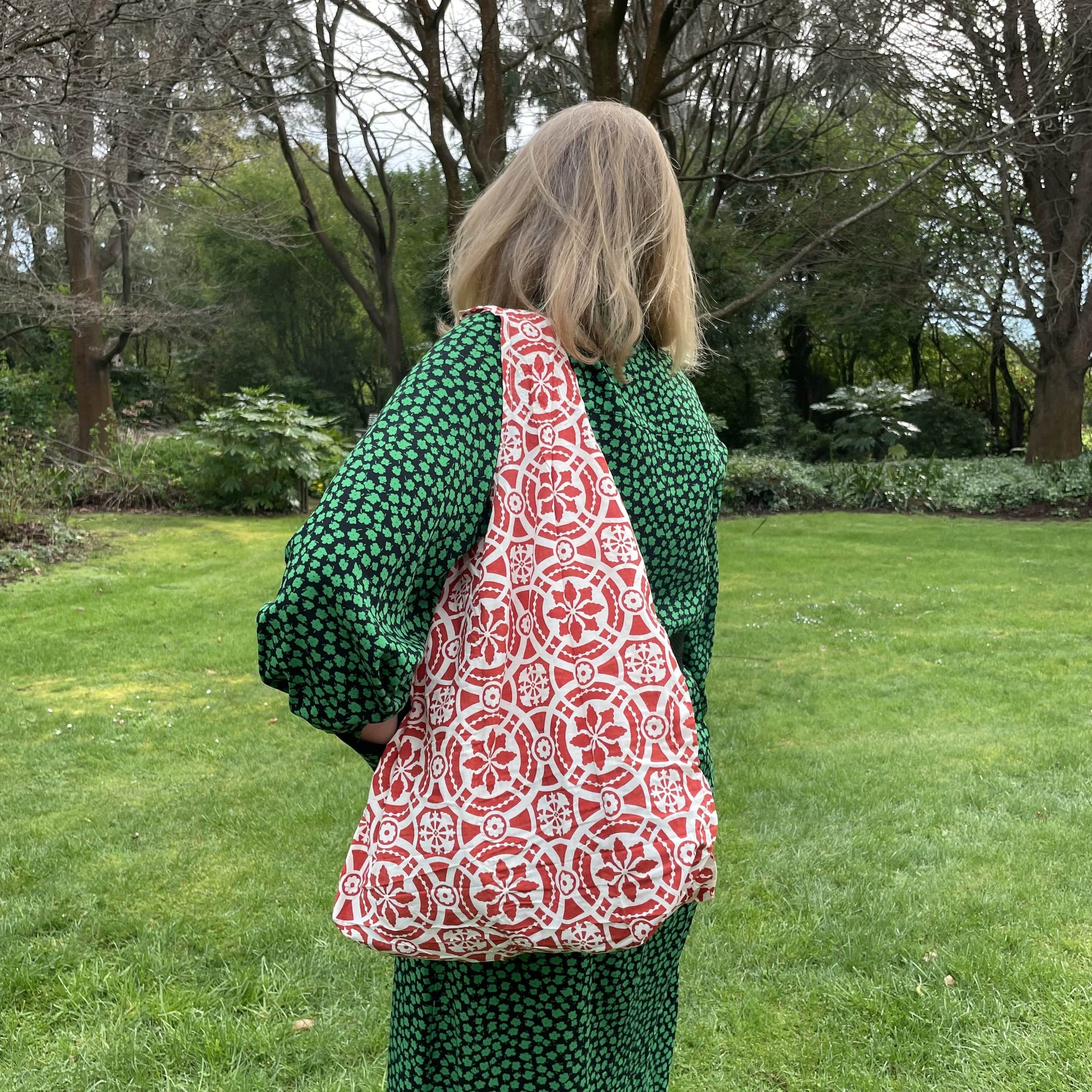 Woman standing in a garden in a black and green dress with her back to the camera. She has a red and white patterned reusable cotton tote bag over her shoulder.