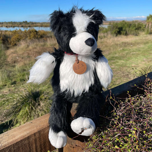 A black & white sheepdog soft toy sitting on a fence overlooking an estuary. The toy dog has a brown leather name tag with REX on it.