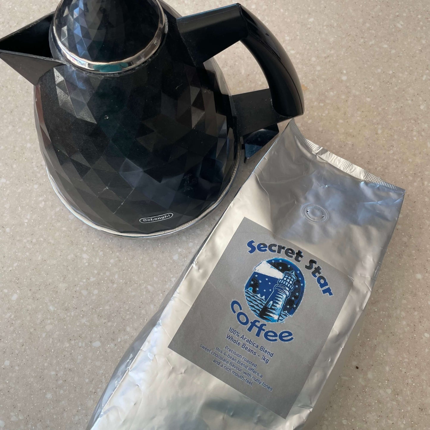Silver bag of coffee beans with label Secret Star Coffee and a black kettle next to it.
