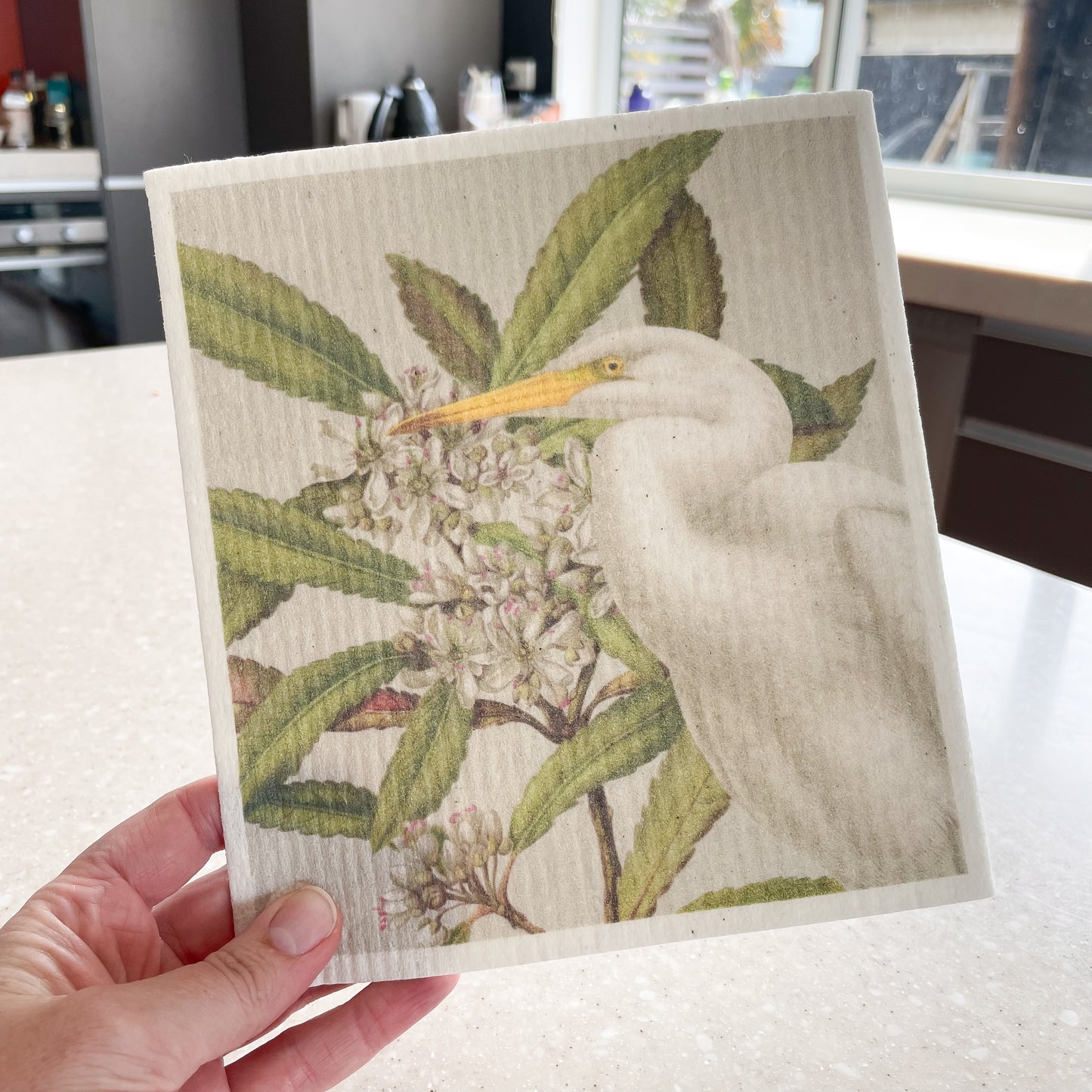 Hand holding a Swedish dishcloth with a kitchen in the background, The dishcloth has a picture of a Kotuku bird and Tawari flowers.