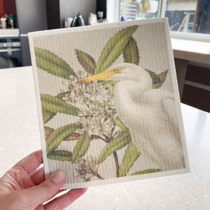 Hand holding a Swedish dishcloth with a kitchen in the background, The dishcloth has a picture of a Kotuku bird and Tawari flowers.