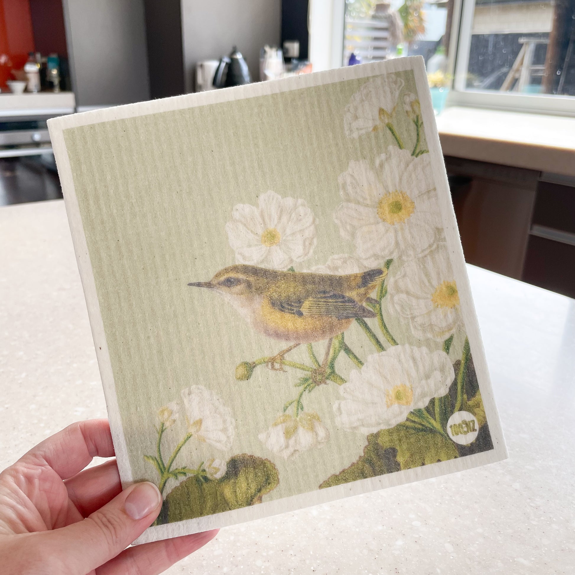 Hand holding a Swedish dishcloth with a kitchen in the background, The dishcloth has a picture of a Rifleman bird and Mt Cook Lilies.