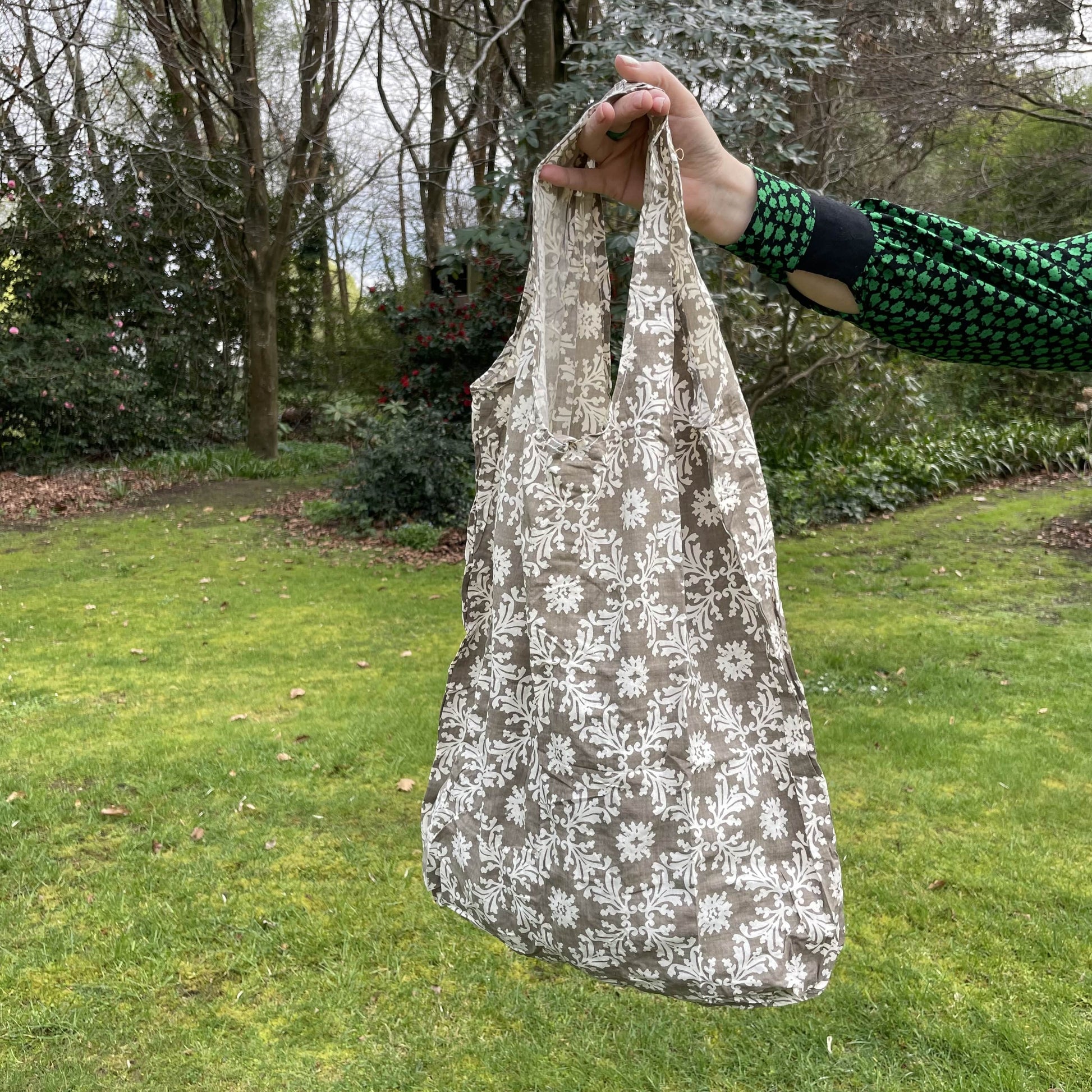 Womans hand holding a floral patterned taupe and white cotton tote bag with a lush green garden in the background.