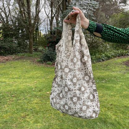 Womans hand holding a floral patterned taupe and white cotton tote bag with a lush green garden in the background.