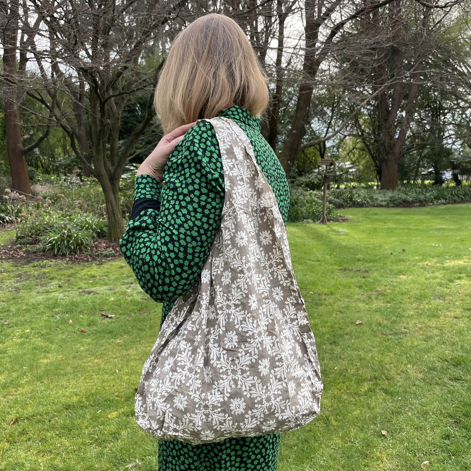 Woman standing in a garden in a black and green dress with her back to the camera. She has a taupe and white floral patterned reusable cotton tote bag over her shoulder.