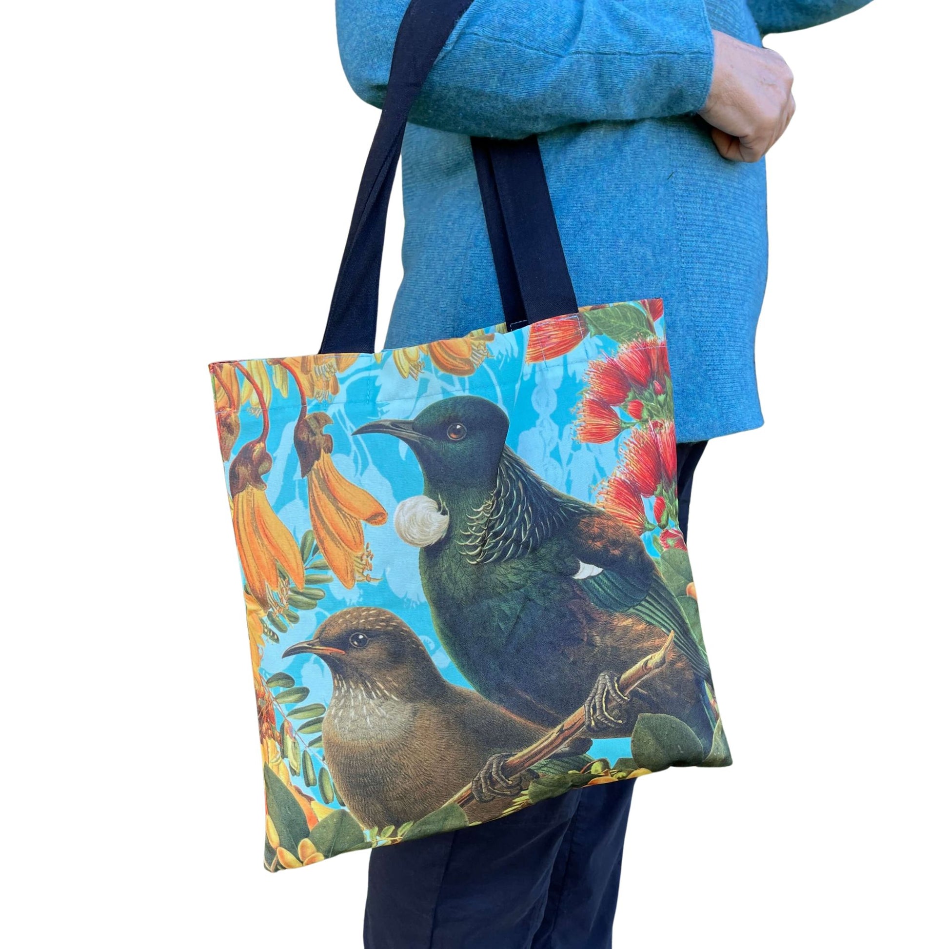 Woman with a tote bag over her arm. The tote features a Bellbird & Tui amongst Kowhai & Pohutukawa flowers.