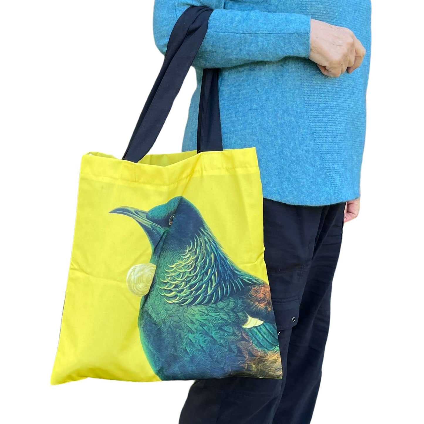Woman with a tote bag over her arm. The bright yellow tote features a Tui bird.