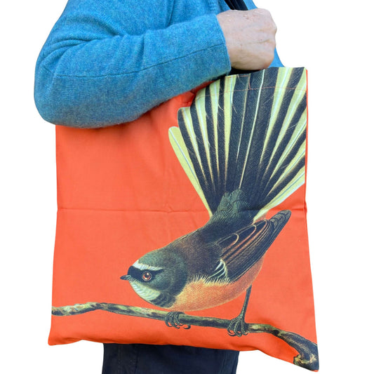 Woman with a tote bag over her arm. The bright orange tote features a fantail bird on a branch.