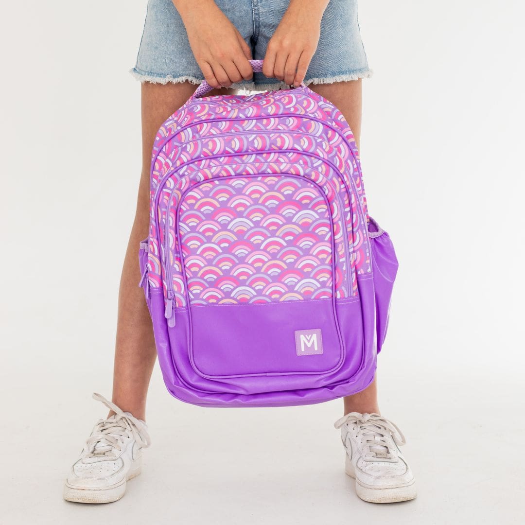 Child holding the top handle of a purple backpack with pink and peach rainbow print by Montiico.