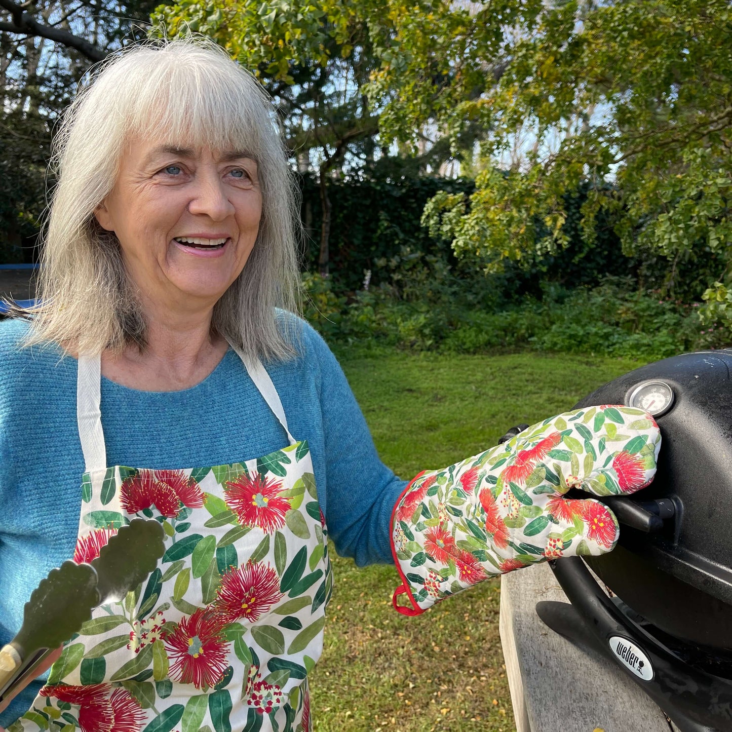 Woman standing by a BBQ wearing a pohutukawa print apron and matching pot mitt.