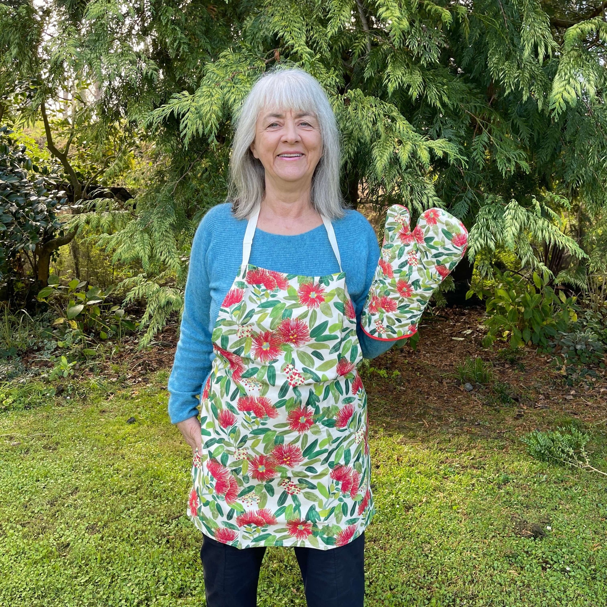 Woman wearing an apron covered in Pohutukawa flower print and showing the matching pot mitt.