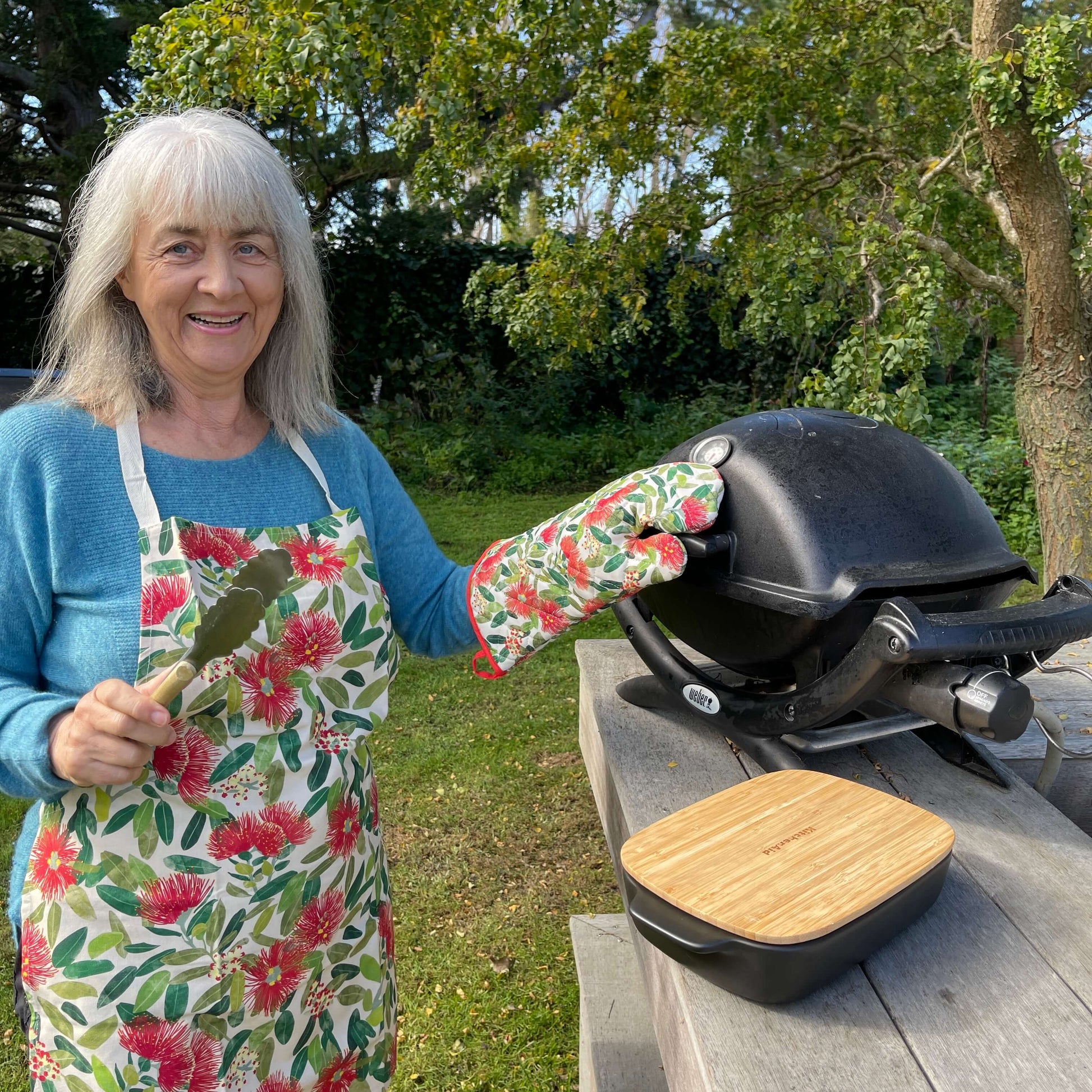 Woman standing by a BBQ wearing an apron covered in Pohutukawa flower print and showing the matching pot mitt.