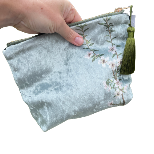Icy blue velvet cosmetic bag with Manuka flowers and a green tassel being held in a hand.