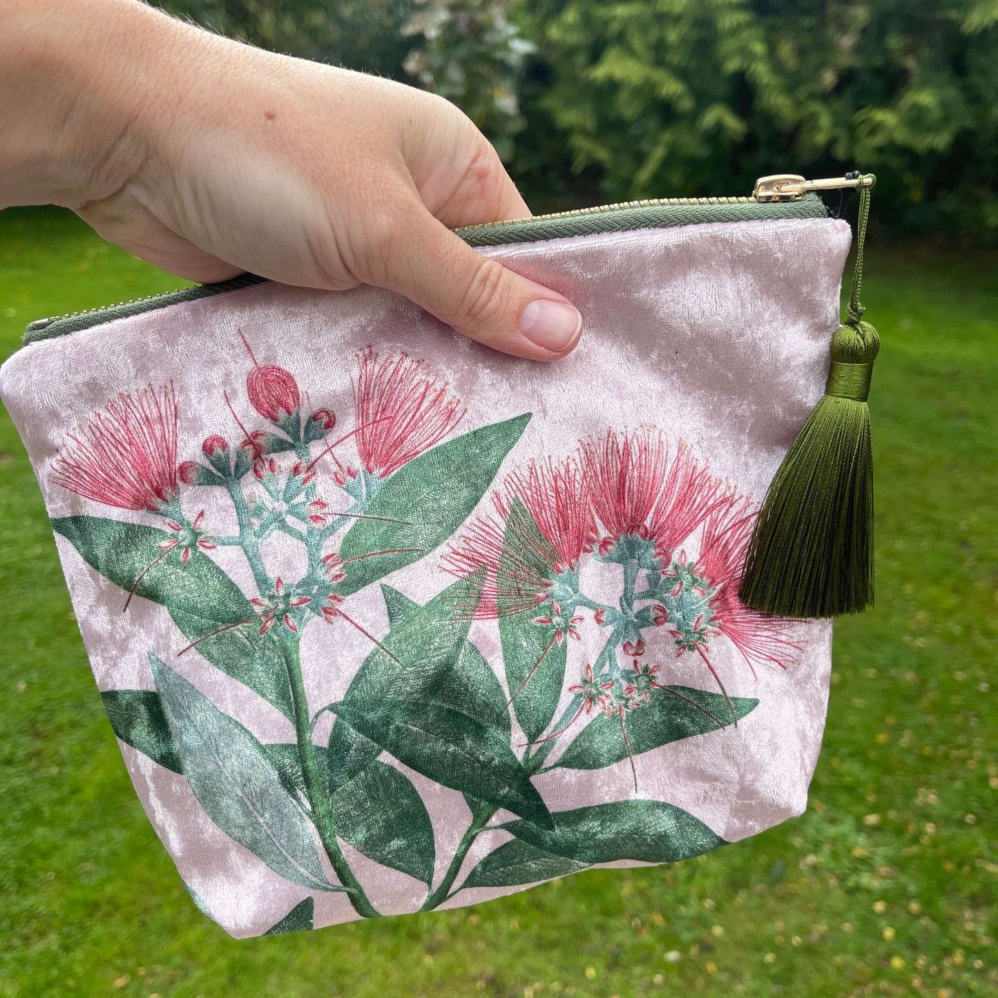 Pale pink velvet cosmetic bag with green tassel and a pohutukawa flower print, being held by a person.
