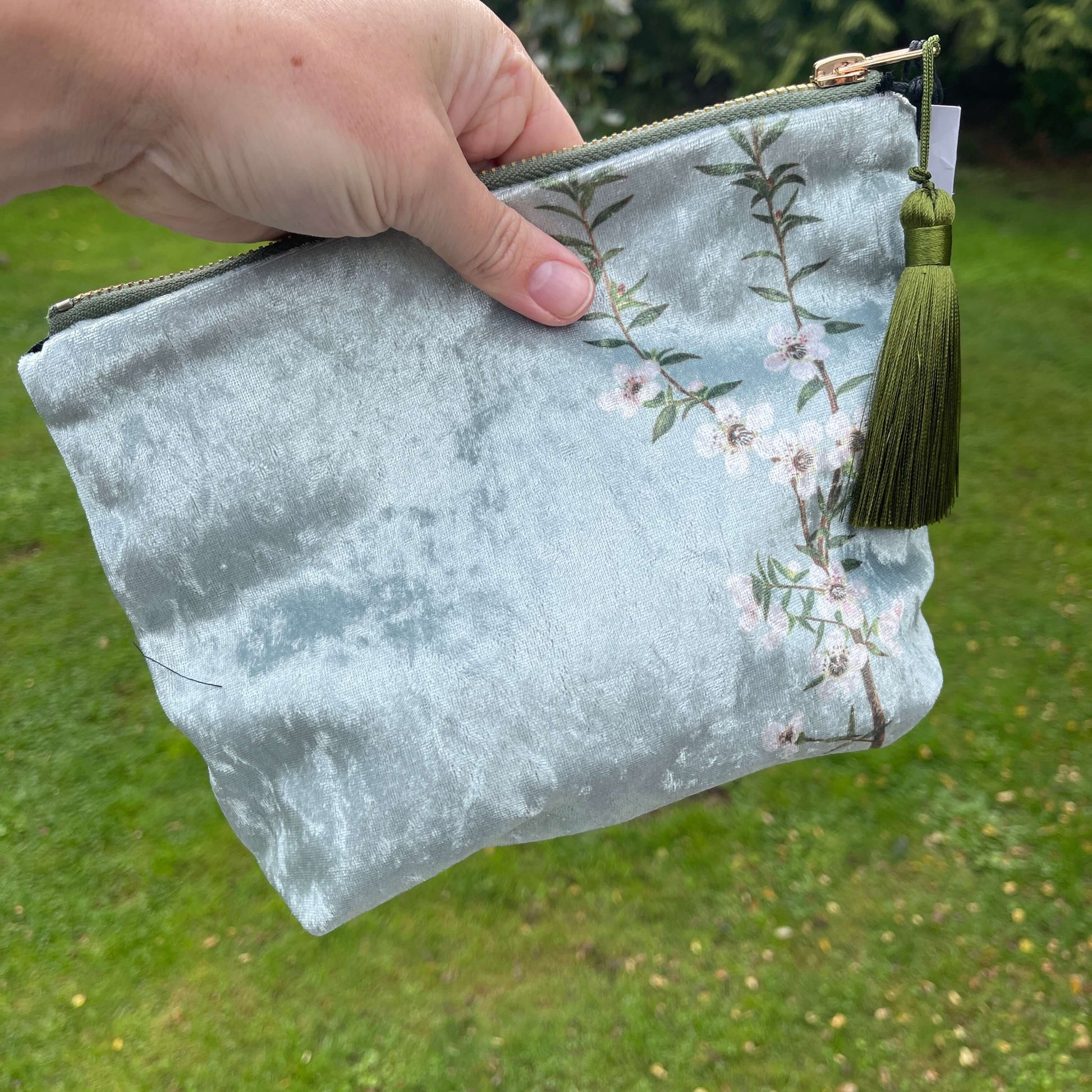 Icy blue velvet cosmetic bag with Manuka flowers and a green tassel being held in a hand.