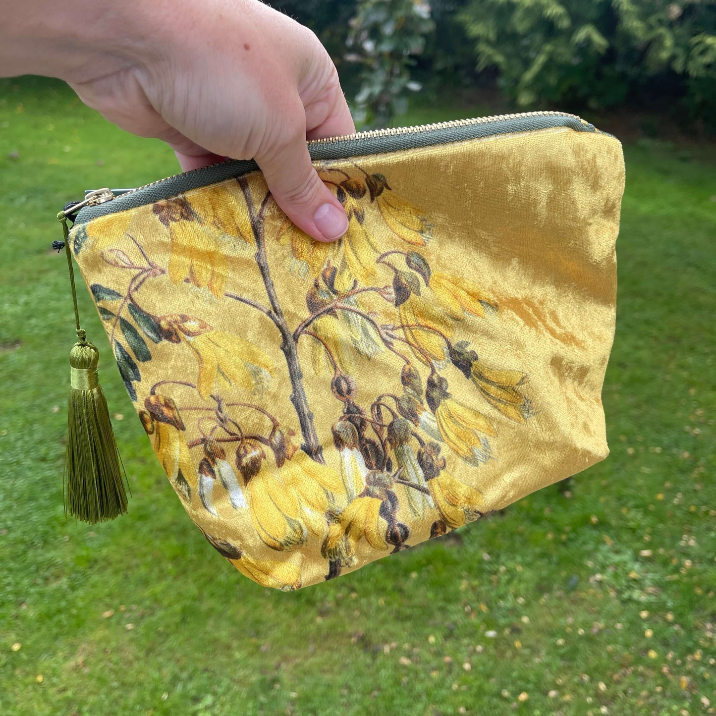 Golden velvet cosmetic bag with Kowhai flowers on it being held in a hand.