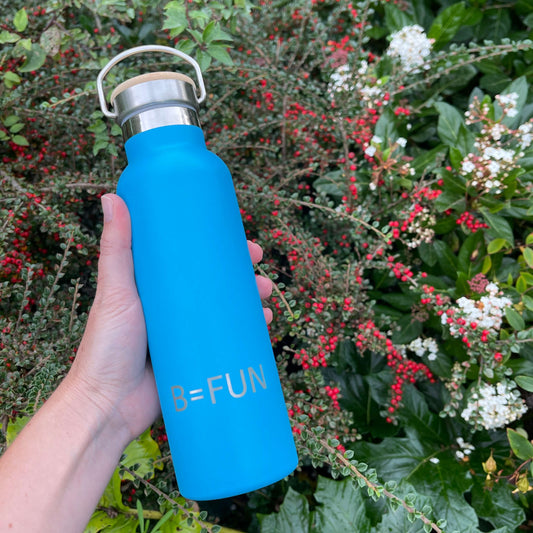 Womens hand holding a bright blue stainless drink bottle with a bamboo cap and the words B=FUN engraved on it.