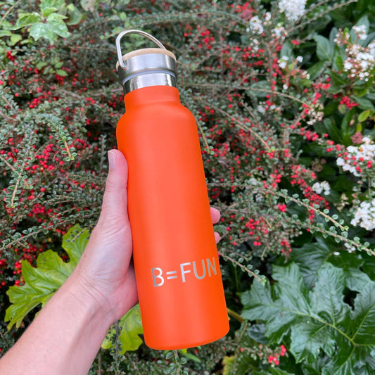 Womens hand holding a bright orange stainless drink bottle with a bamboo cap and the words B=FUN engraved on it.