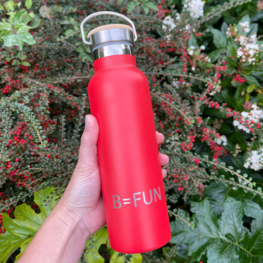 Womens hand holding a bright red stainless drink bottle with a bamboo cap and the words B=FUN engraved on it.