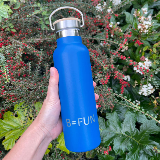Womens hand holding a bright blue stainless drink bottle with a bamboo cap and the words B=FUN engraved on it.