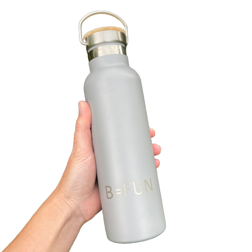 Womens hand holding a stormy grey stainless drink bottle with a bamboo cap and the words B=FUN engraved on it.