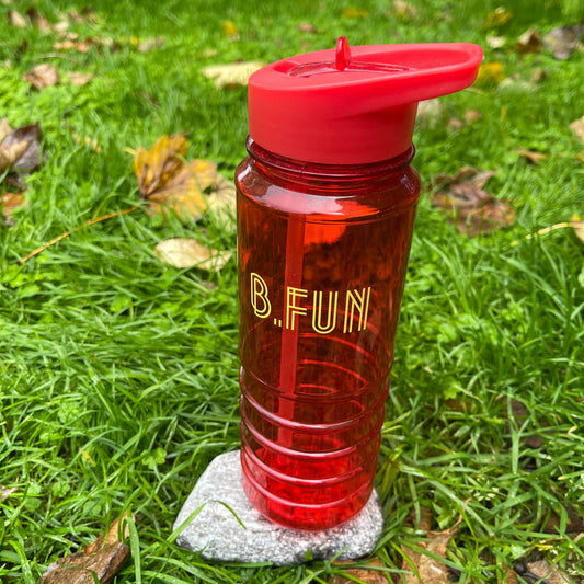 Bright red plastic sports water bottle with B=Fun printed on it sitting on a rock in the grass.