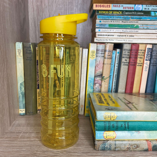 Bright yellow plastic sports water bottle with B=Fun printed on it sitting on a book shelf next to vintage books.