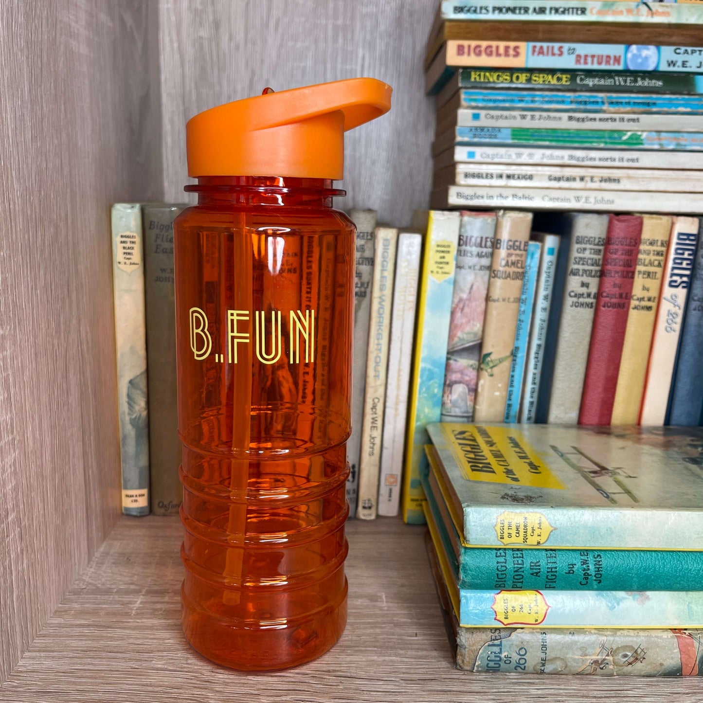Bright orange plastic sports water bottle with B=Fun printed on it sitting on a book shelf next to vintage books.