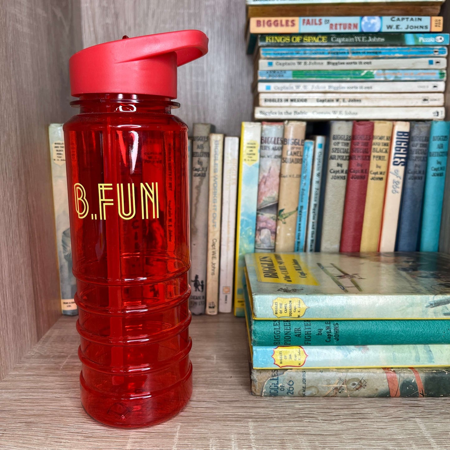 Bright red plastic sports water bottle with B=Fun printed on it sitting on a book shelf next to vintage books.
