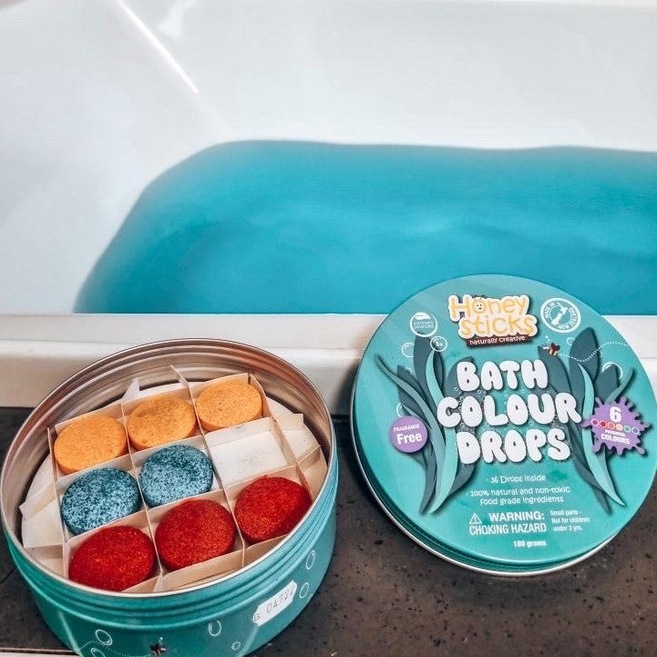 Honey Sticks bath colour drops in a tin next to a bath with blue water.