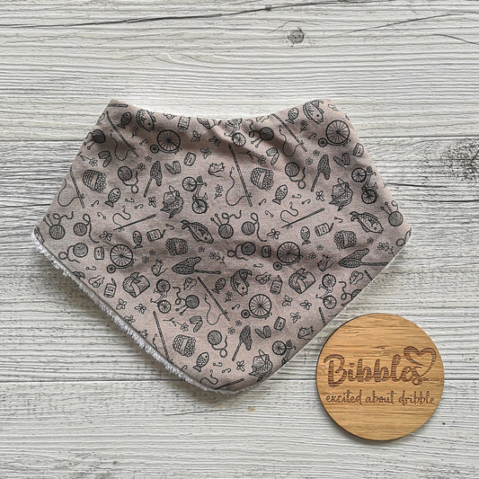 Baby dribble bib in a light mushroom colour with fish, yarn, penny farthings and other little icons in a darker brown print.